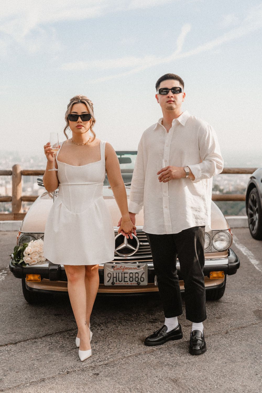 Stylish couple in white outfits pose in front of vintage Mercedes car during sunset photoshoot.