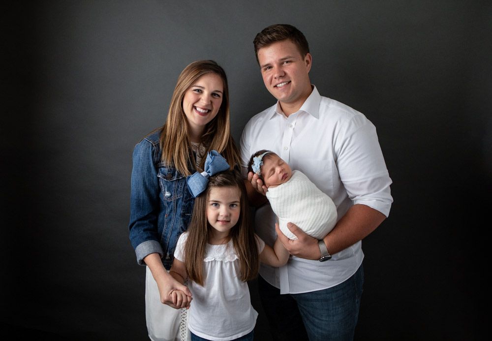 Family portrait with newborn in casual attire against dark background.