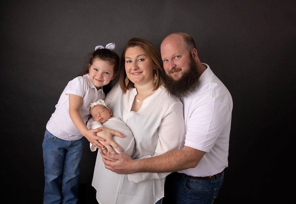 Studio portrait of a family of four with newborn baby against dark backdrop.