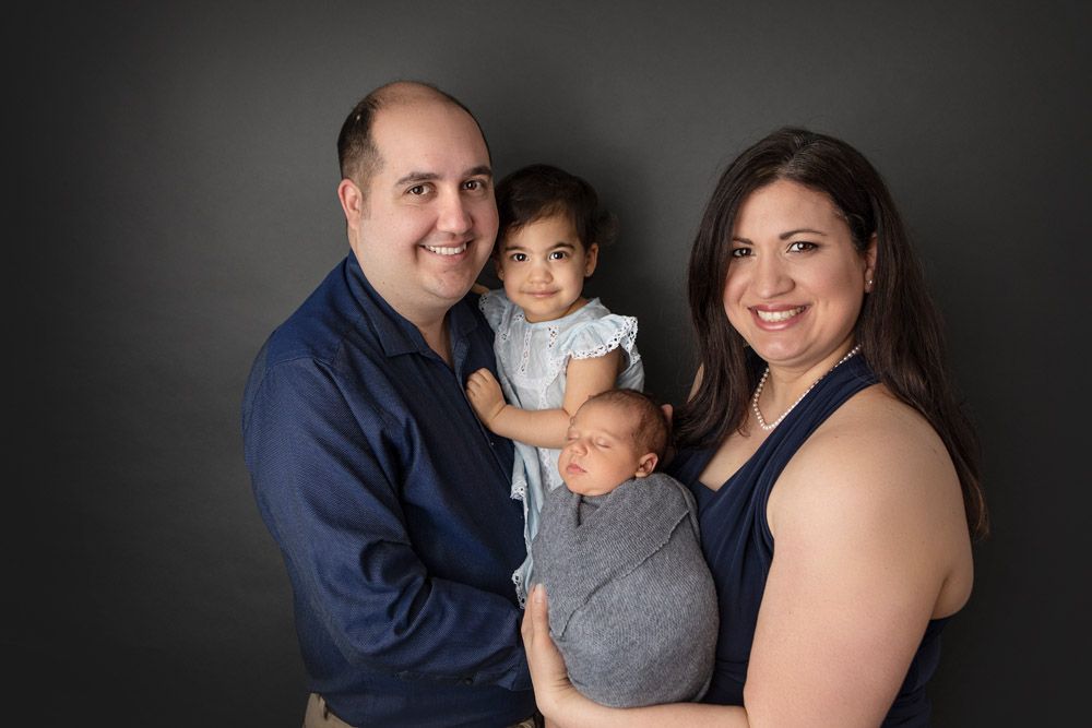Studio family portrait with toddler and newborn against dark background.
