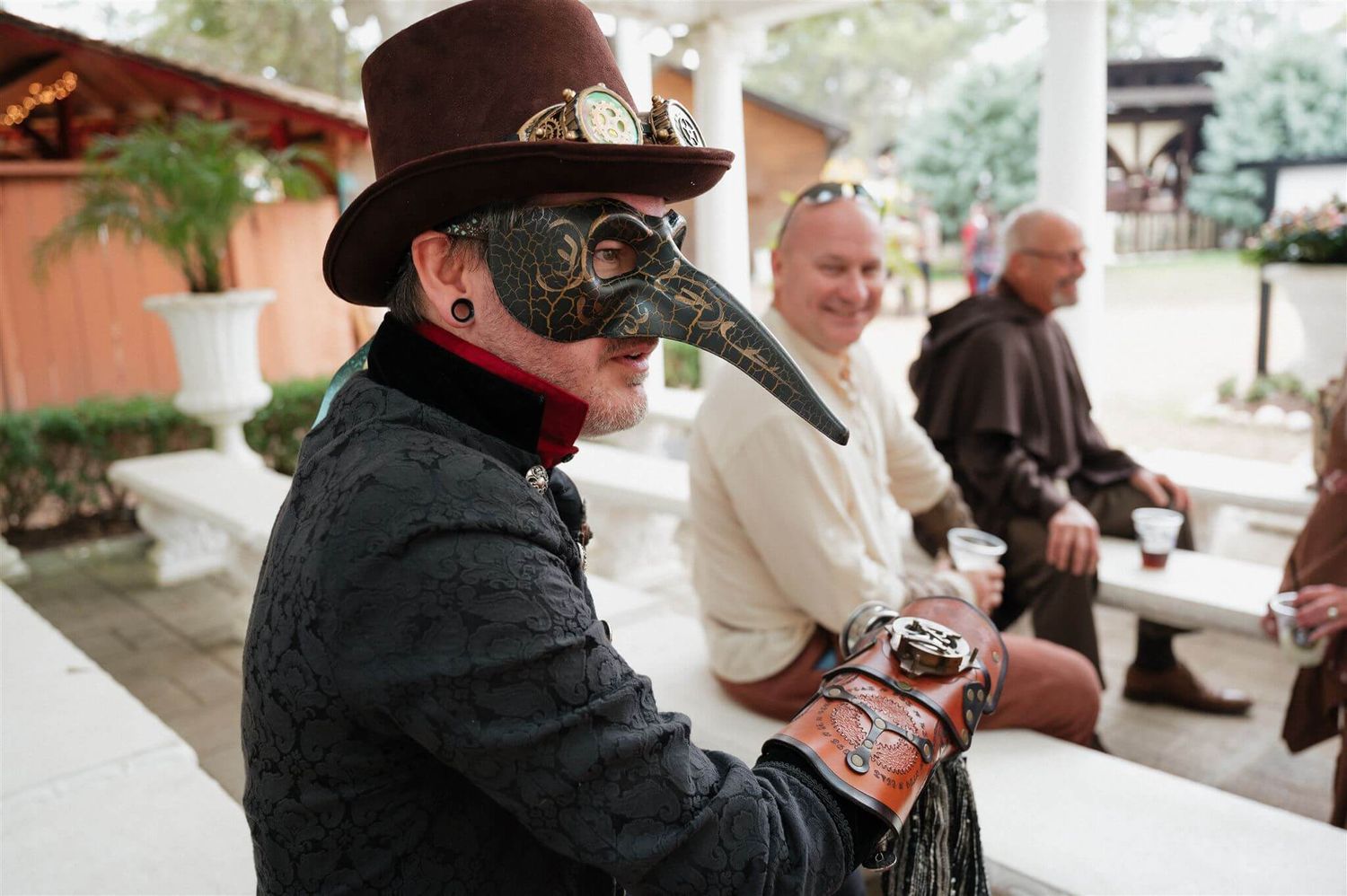 Guest in plague doctor mask and period costume seated at outdoor renaissance wedding celebration.