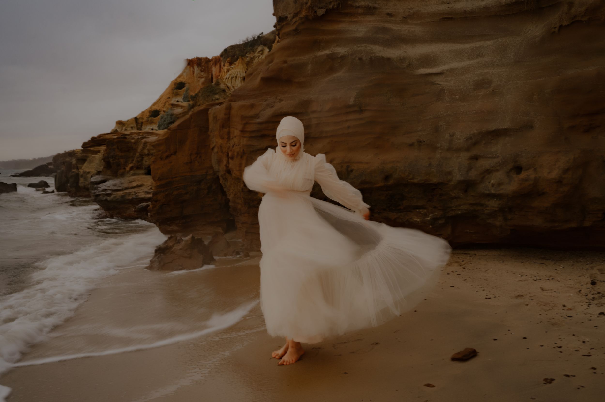 A flowing white dress catches the ocean breeze against dramatic coastal cliffs during a moody beach photoshoot.
