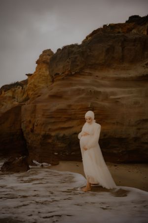 An ethereal white gown contrasts against rugged sandstone cliffs at a beach during golden hour.