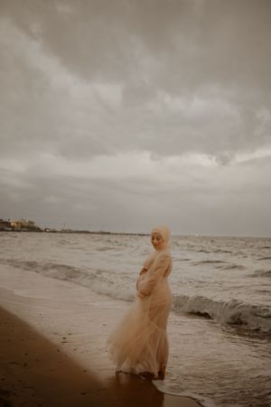 A dreamy coastal maternity shoot features flowing white fabric against stormy skies and crashing waves.
