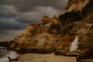 Dramatic coastal cliffs rise above a sandy beach under moody storm clouds at sunset.