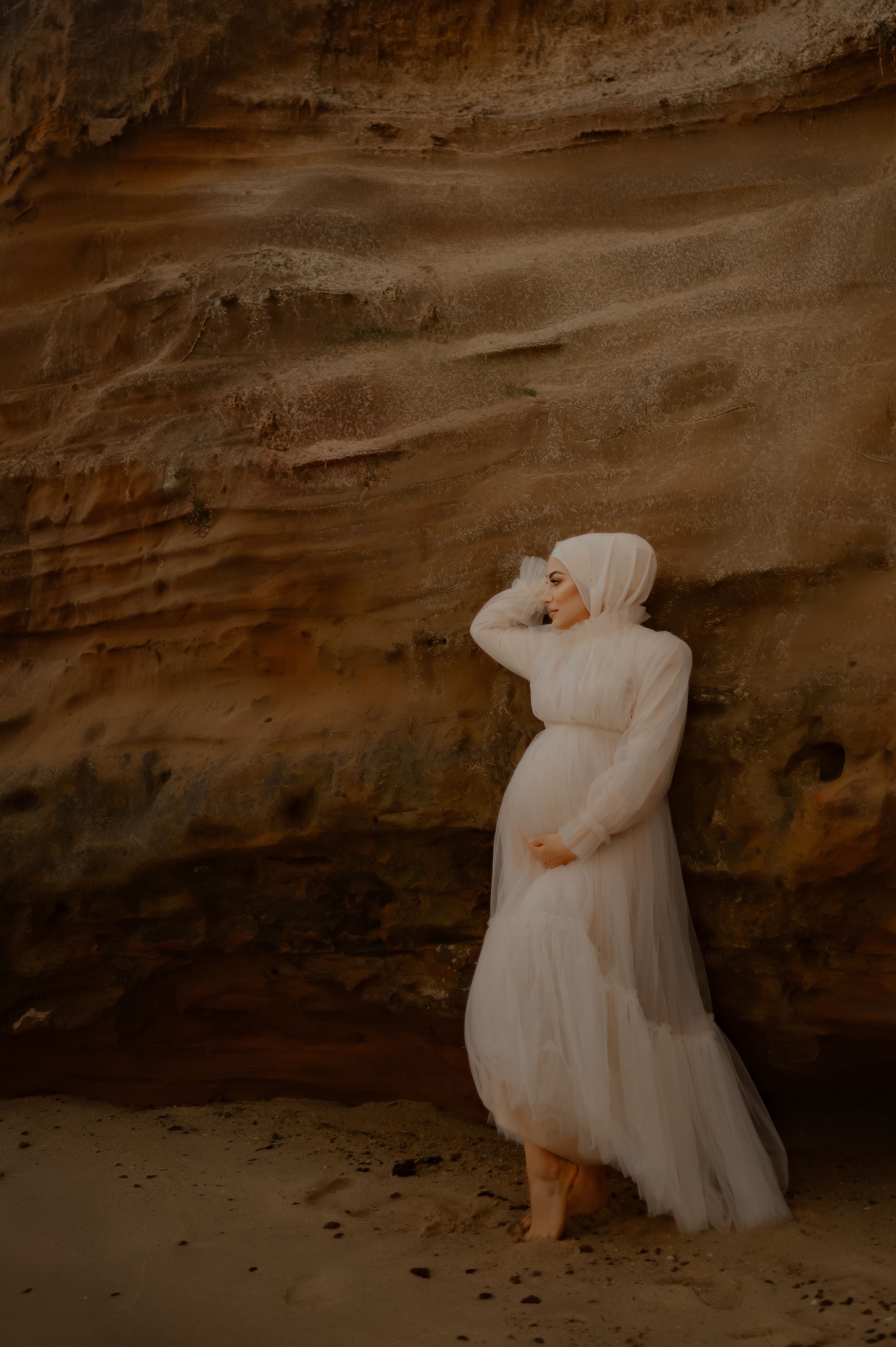 A romantic maternity photoshoot features flowing white fabric against weathered coastal cliffs.