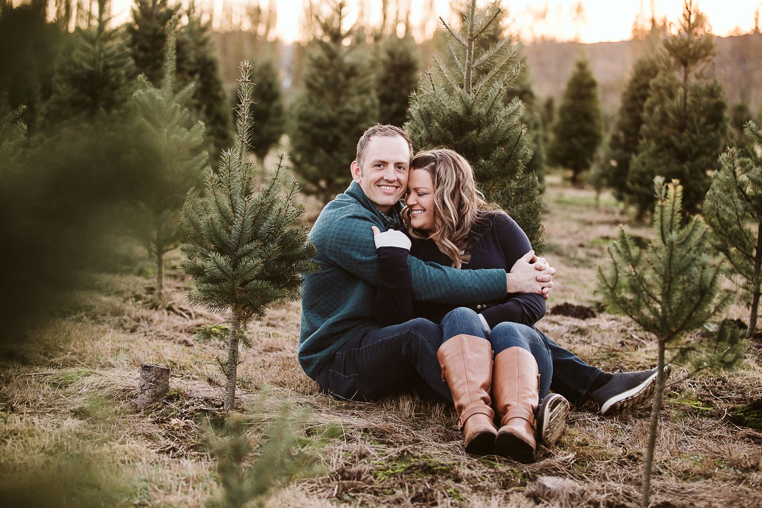A couple cuddles on the ground among young evergreen trees at sunset.