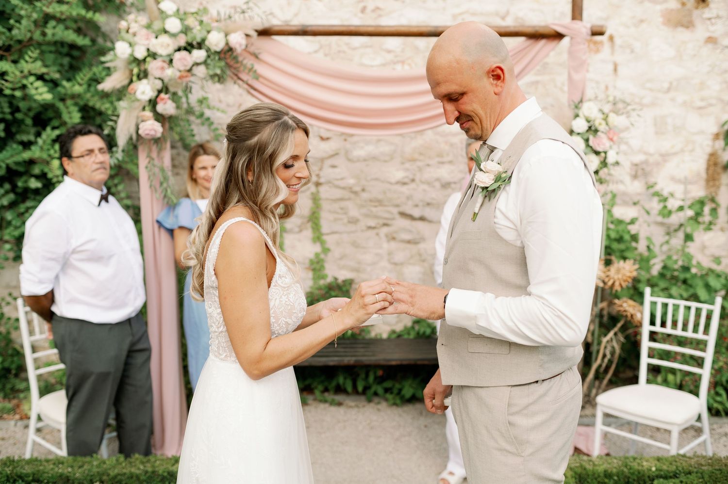 Wedding couple exchanges rings during outdoor ceremony with blush drapery and stone wall background.