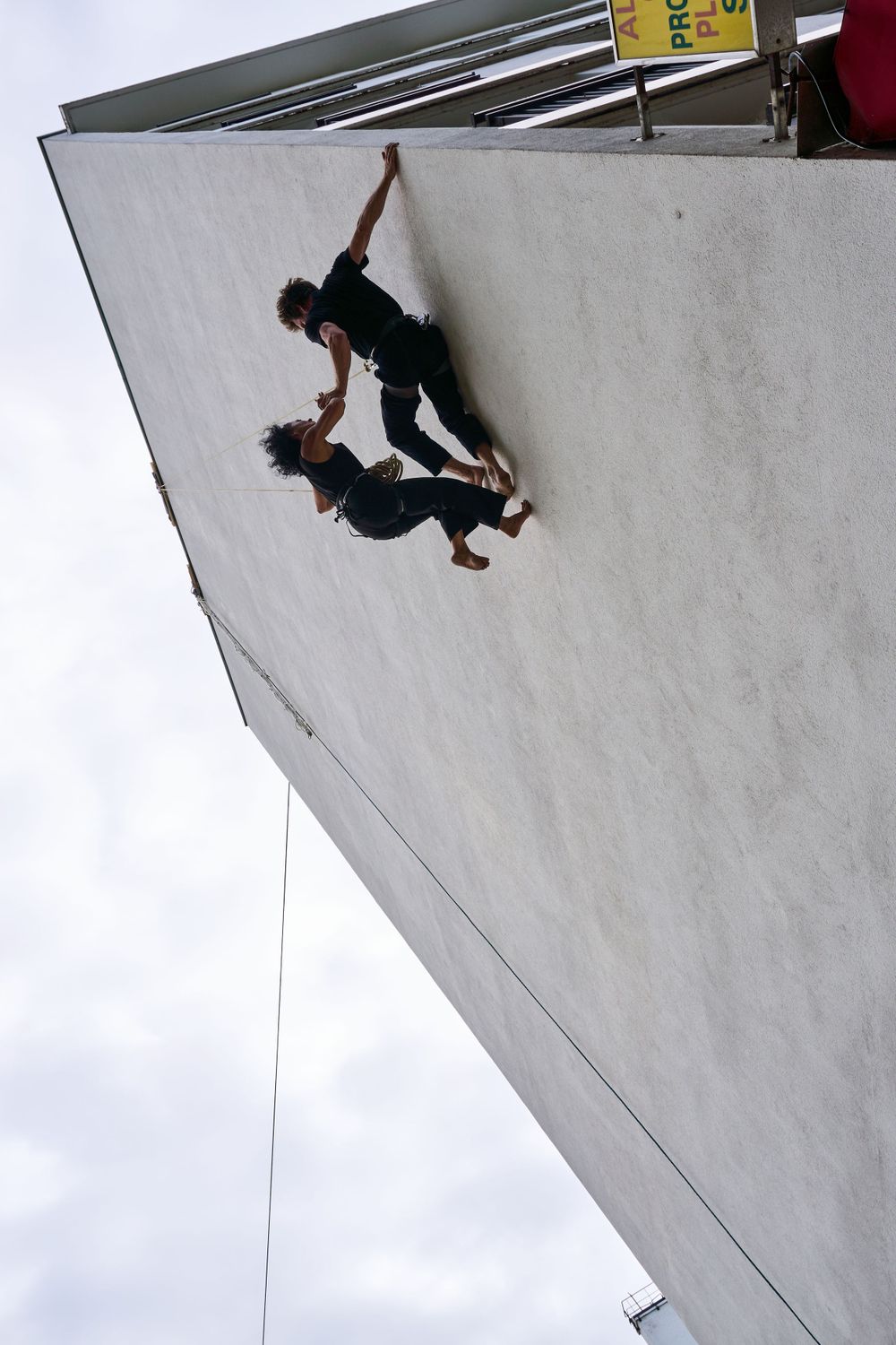 Two performers execute a dramatic aerial dance move on a steep wall surface.