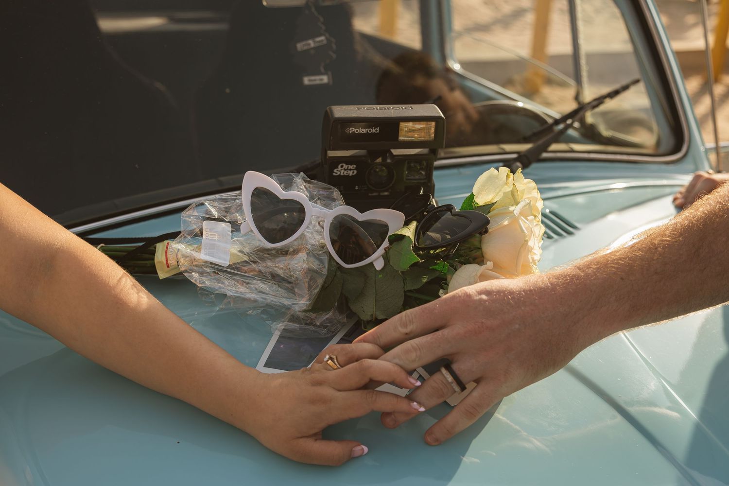 Close-up of hands with rings next to heart sunglasses and flowers on vintage blue car.