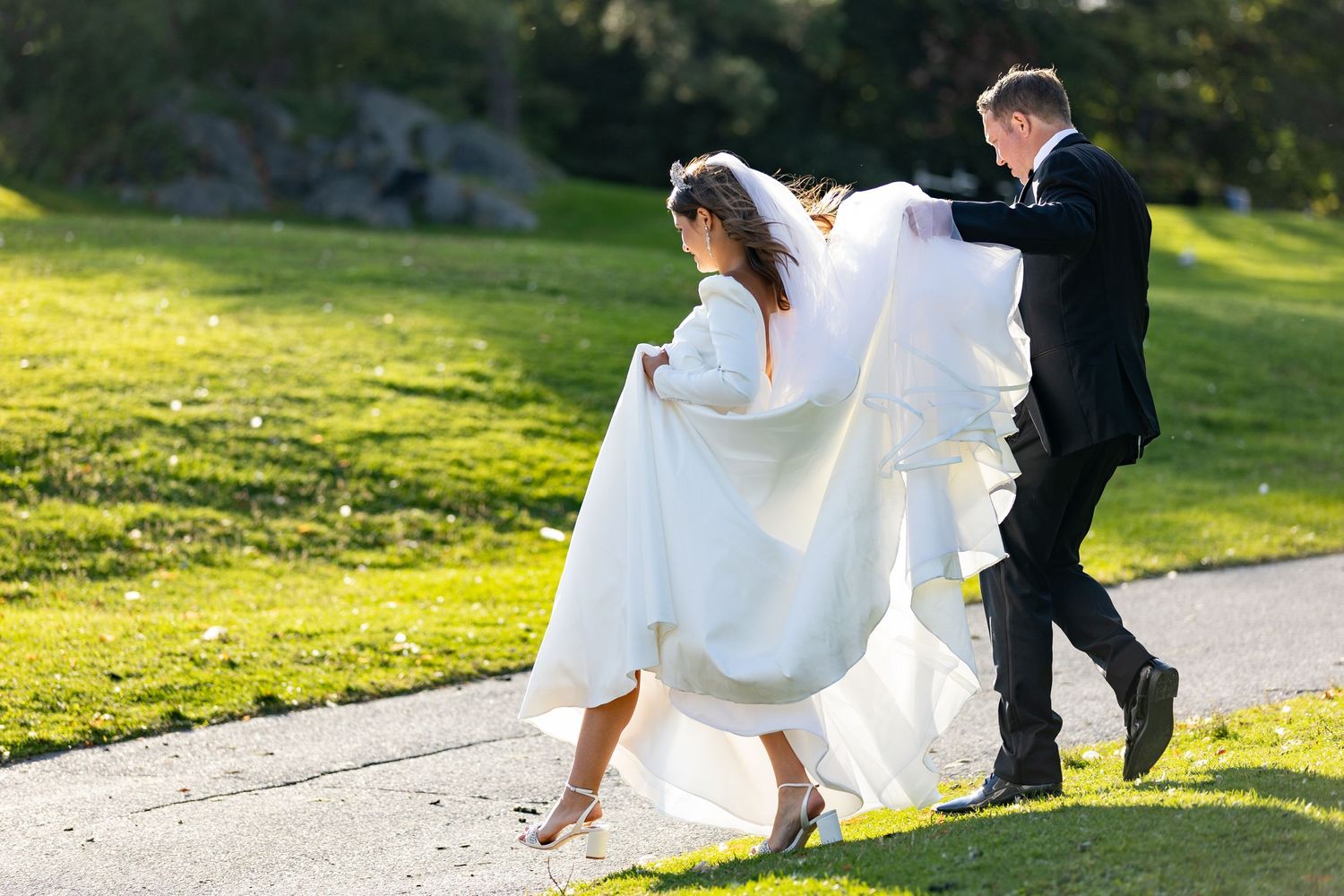 Bride and groom walk down green hill toward paved path, groom holding bride's train.