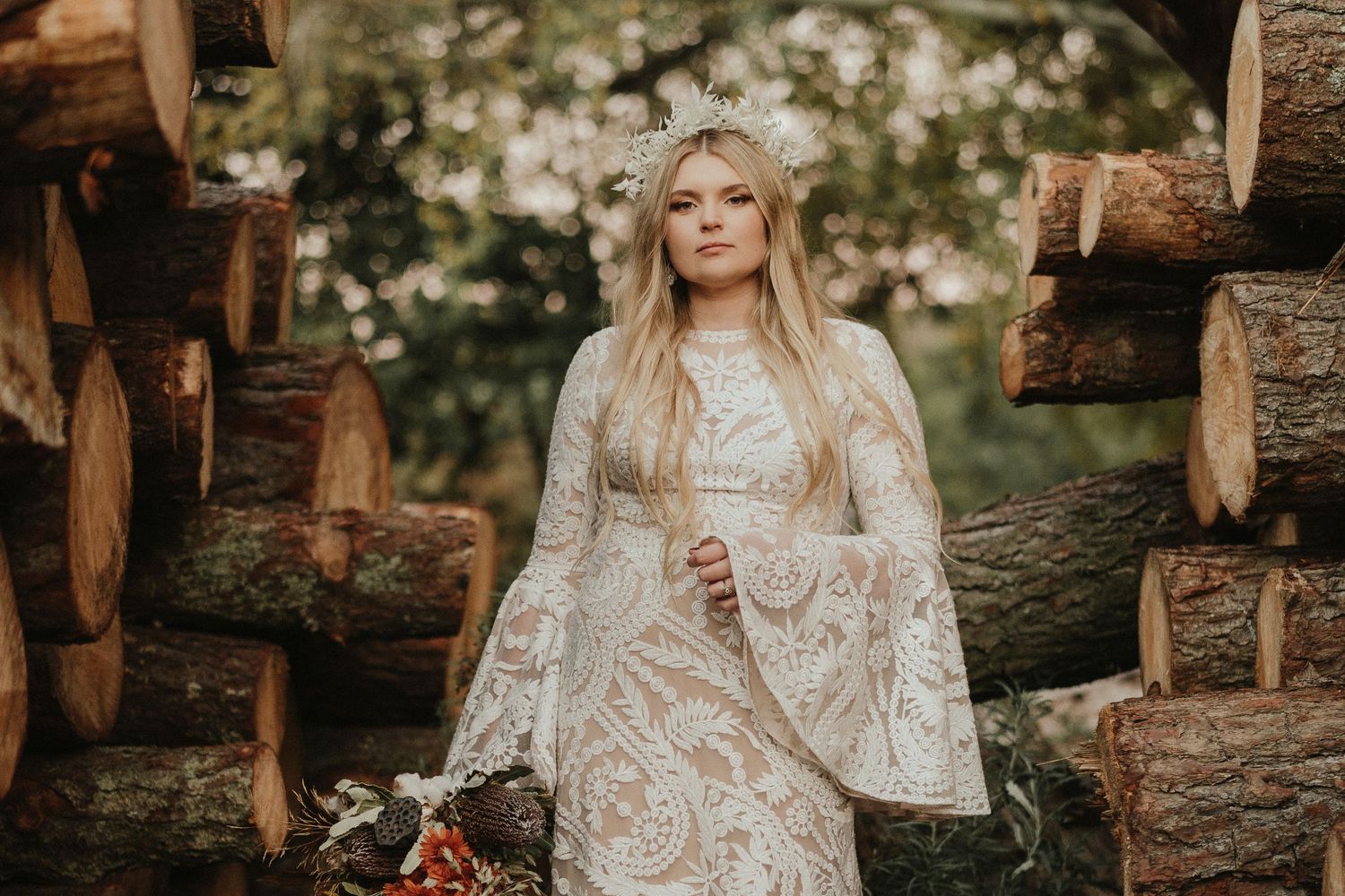 Boho bride in flowing lace dress stands between rustic wood piles.