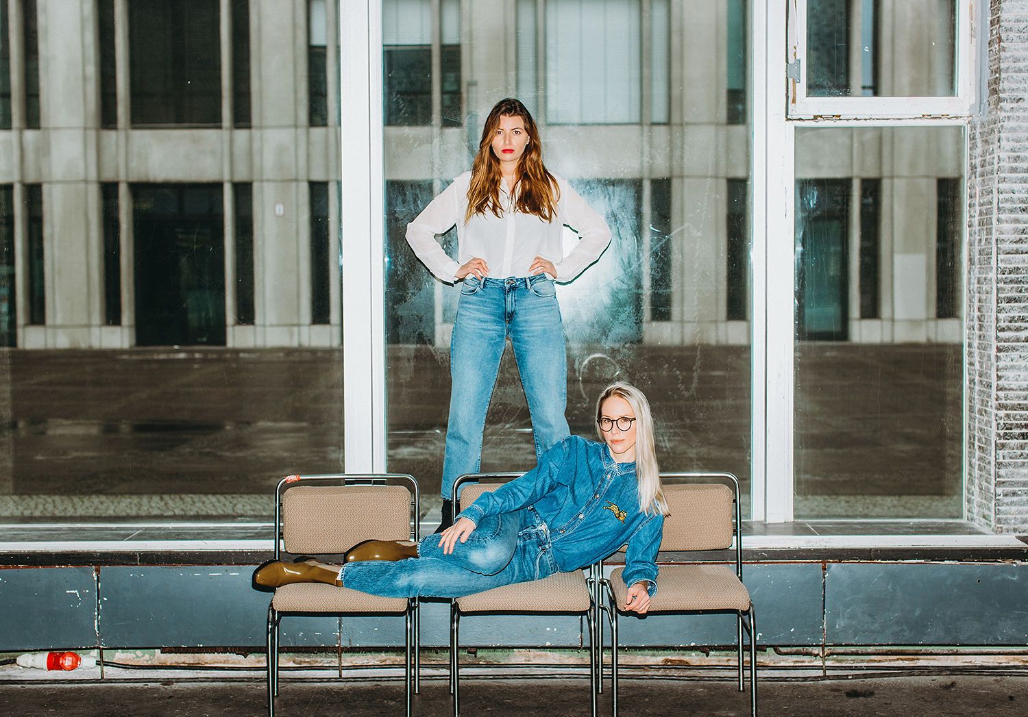 Two people in casual outfits posing playfully on metal benches in modern building.
