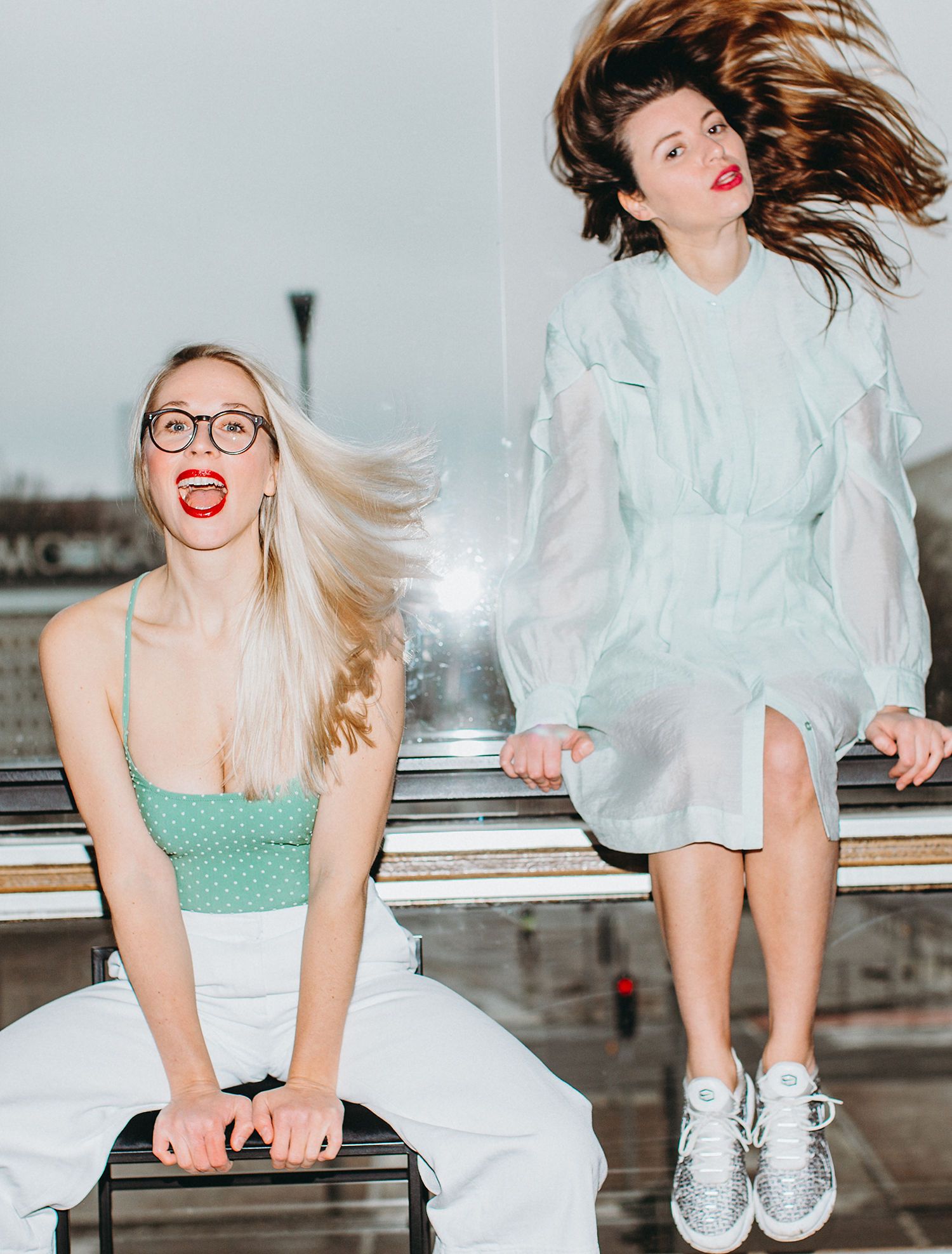 Two models with flowing hair pose energetically on urban rooftop in contrasting outfits.