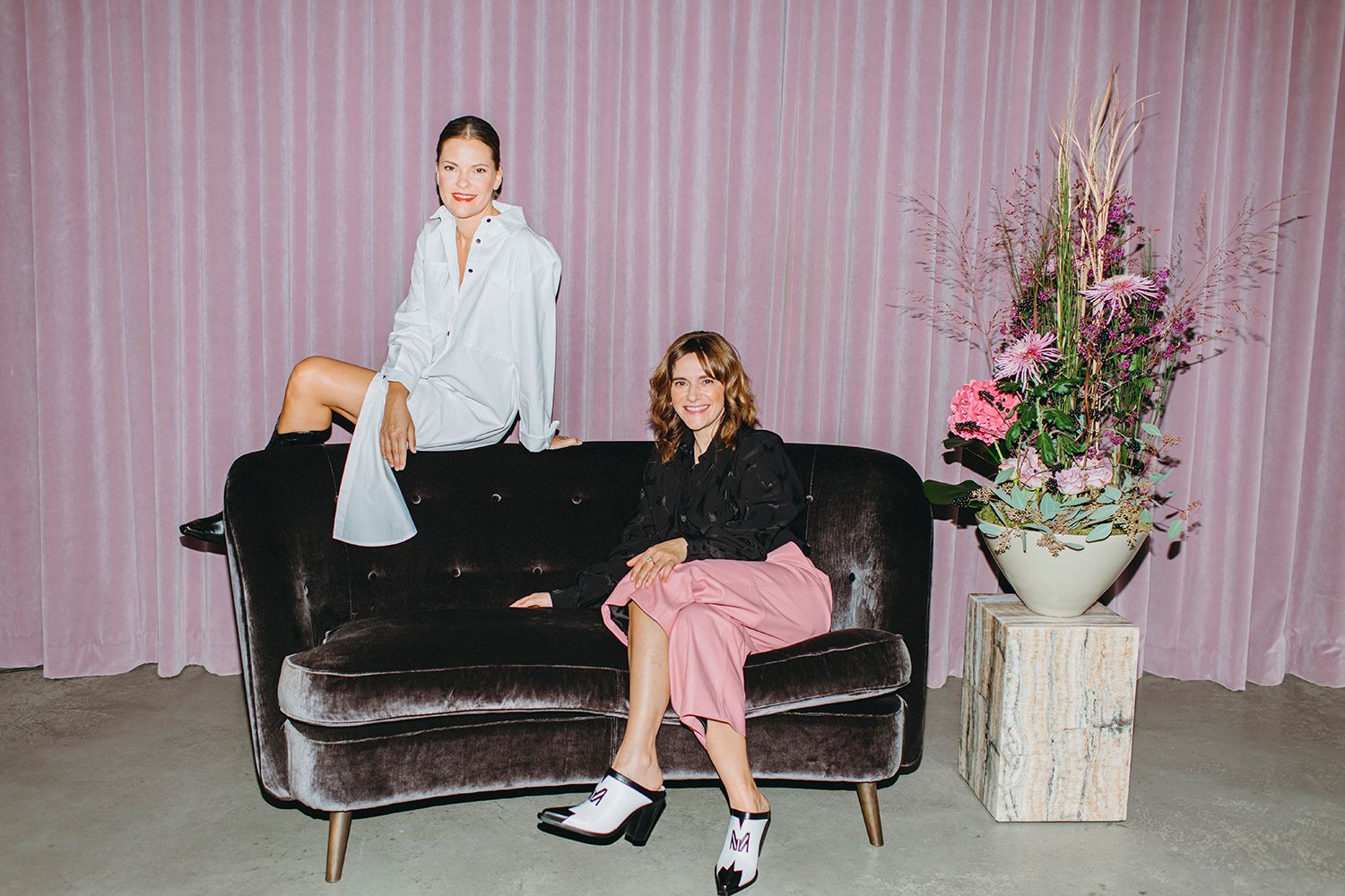 Two people lounging casually on velvet couch with pink curtain and floral arrangement.