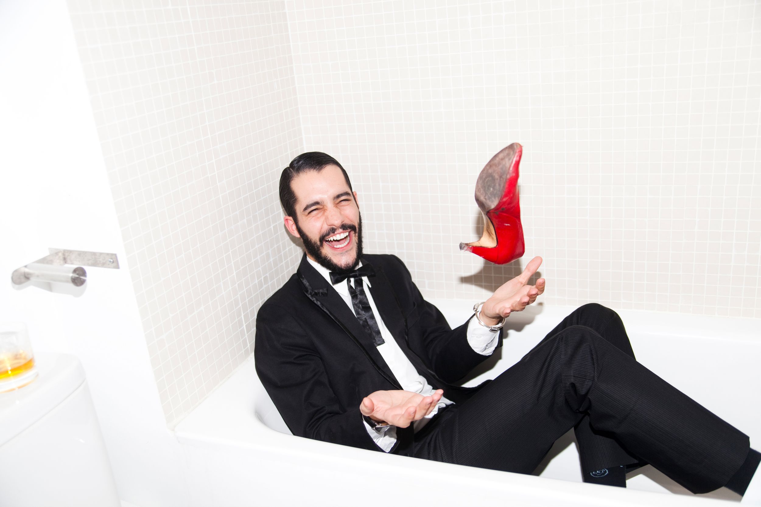 Person in black suit lying on floor holding red high heel shoe.