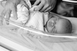 Black and white photo of adult hands gently swaddling peaceful newborn baby in hospital bassinet.