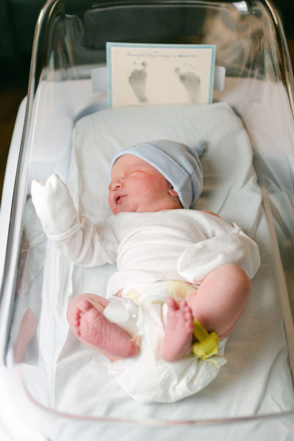 Newborn baby in white onesie and blue hat lying in hospital bassinet with footprint card nearby.
