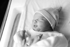 Black and white close-up of sleeping newborn baby wearing white knit hat in hospital bassinet.