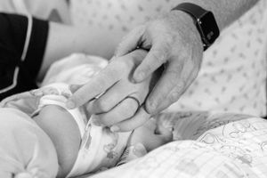 Black and white close-up of father holding tiny newborn baby fingers in hospital setting.