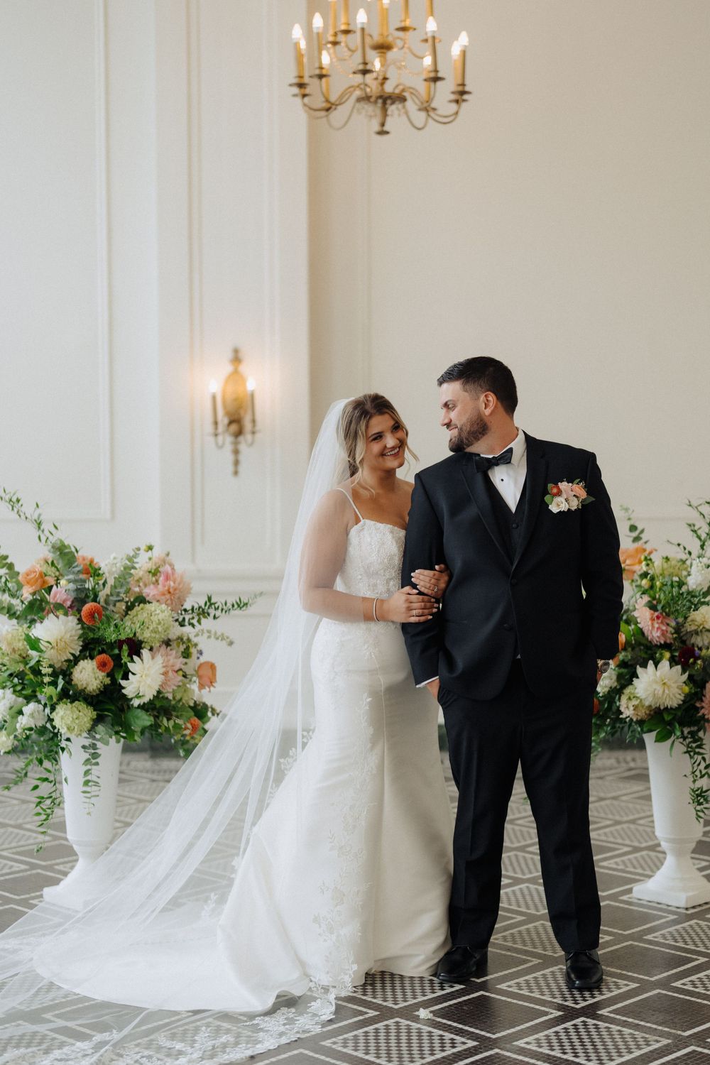 Elegant couple poses in front of floral arrangement with chandelier in classic wedding venue.