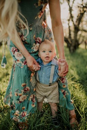 Young child in suspenders stands in grass while held by someone wearing a floral dress.