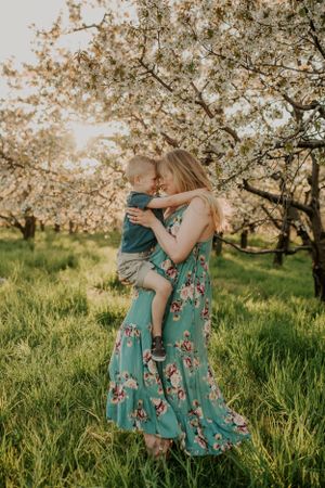 Person in floral dress holds child among blooming cherry trees in spring.