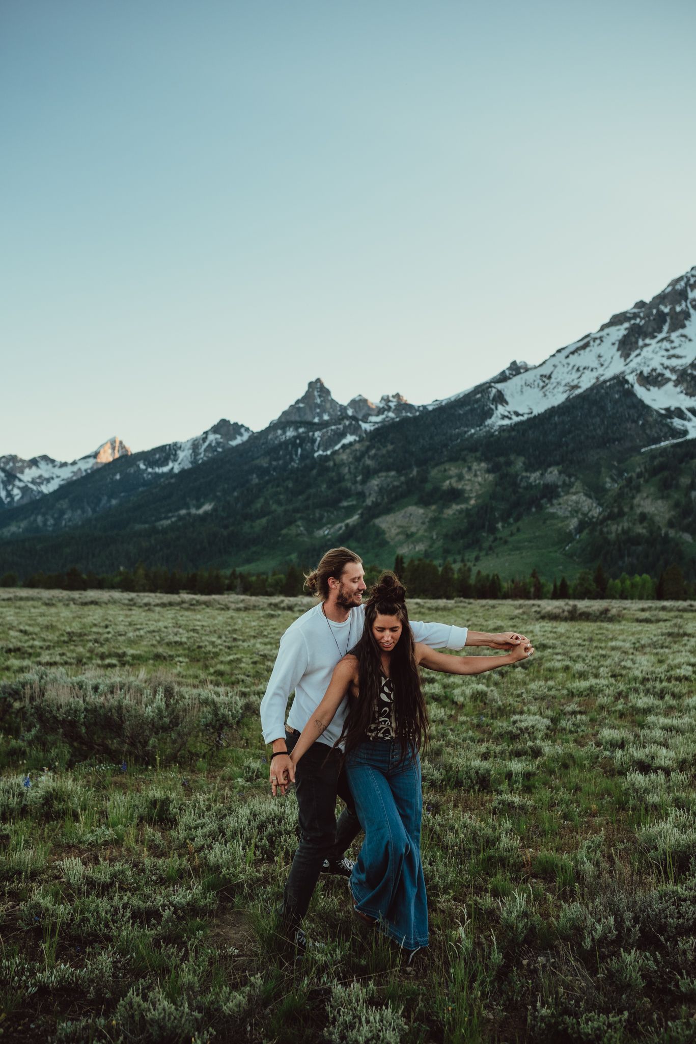 A couple enjoying a romantic moment in a meadow with snow-capped mountains in the background at dusk.