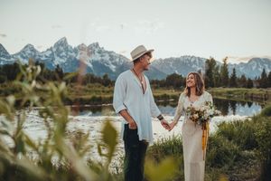 Couple in casual attire standing by mountain lake with wildflower bouquet.