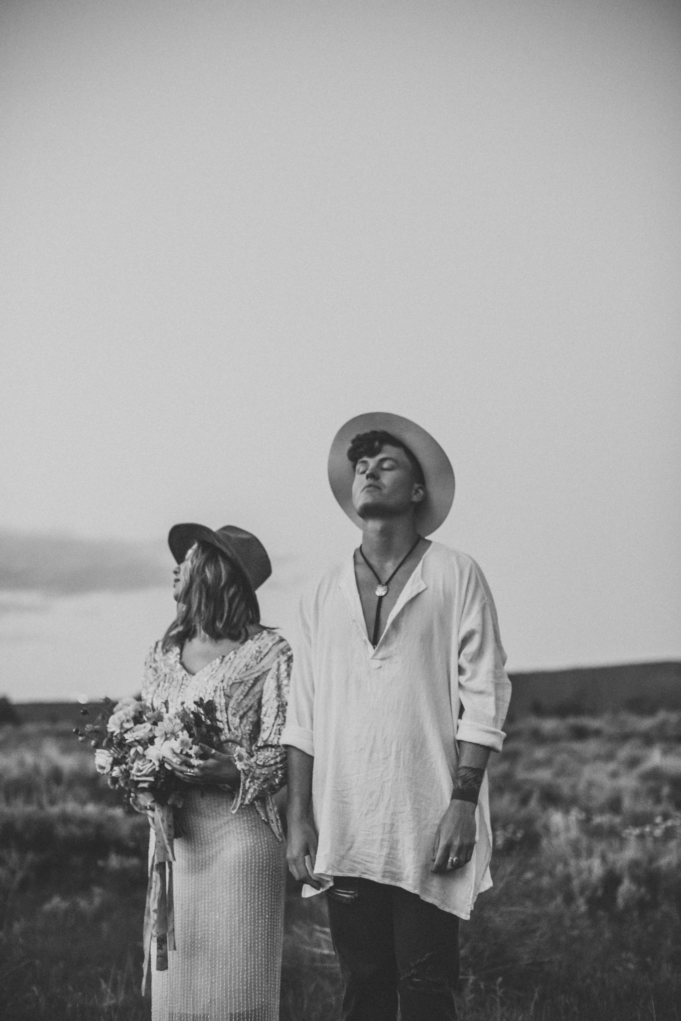 Artistic black and white portrait of couple in hats against misty landscape.