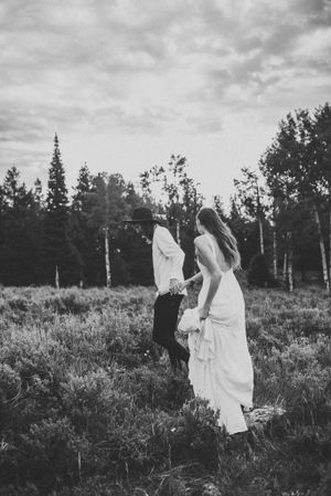Black and white photo of couple dancing in field with forest backdrop and cloudy sky.