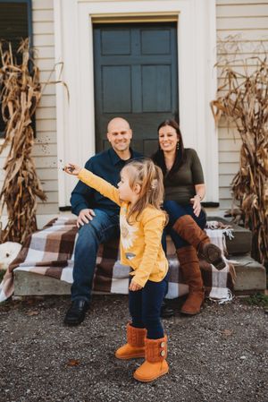 Family sits on porch steps with fall decorations while child in yellow jacket plays.