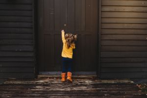 Child in yellow coat and boots plays in front of dark wooden door.