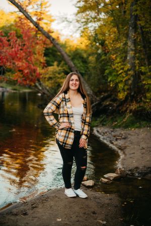 Person in plaid jacket stands by riverside with colorful fall trees in background.