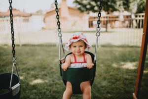 Toddler wearing red hat smiles while swinging in backyard playground.