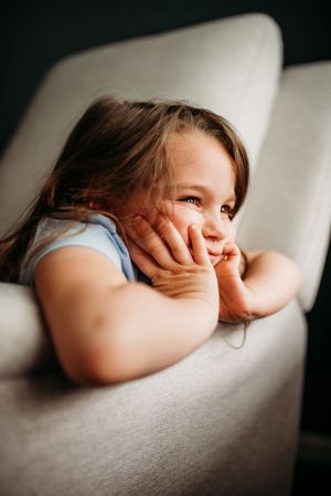 Young child resting on a white couch with hands on cheeks, smiling and looking off to the side.