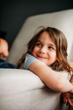 Child laying on white couch with playful expression and hands tucked under chin.