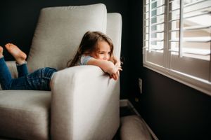 Child relaxing on light colored armchair near window with plantation shutters.