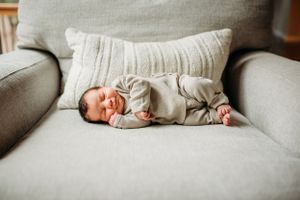 Sleeping newborn in beige onesie lying on gray couch with decorative pillow.