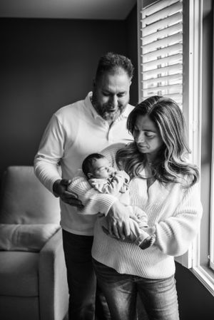 Family with newborn baby sharing tender moment by window in black and white photograph.