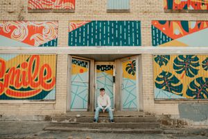 Person sitting in front of colorful street art mural on brick building with geometric patterns.