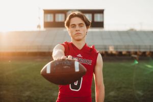 Football player in red jersey poses with ball on field at sunset.
