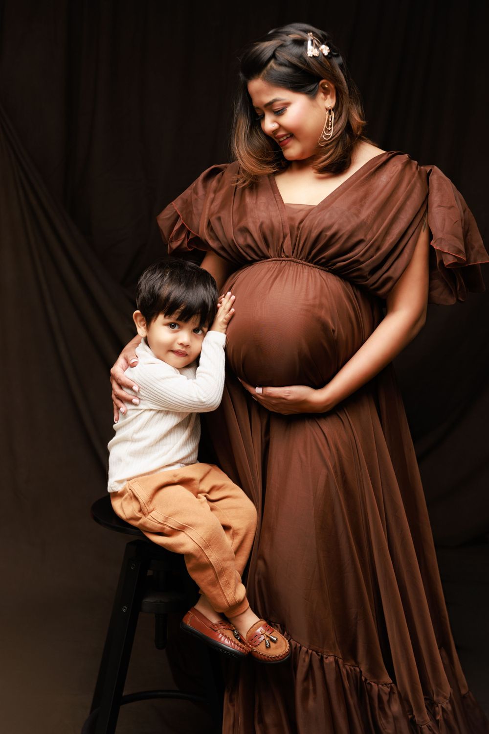 A touching maternity portrait of a pregnant woman in brown dress embracing her young child against a dark backdrop.