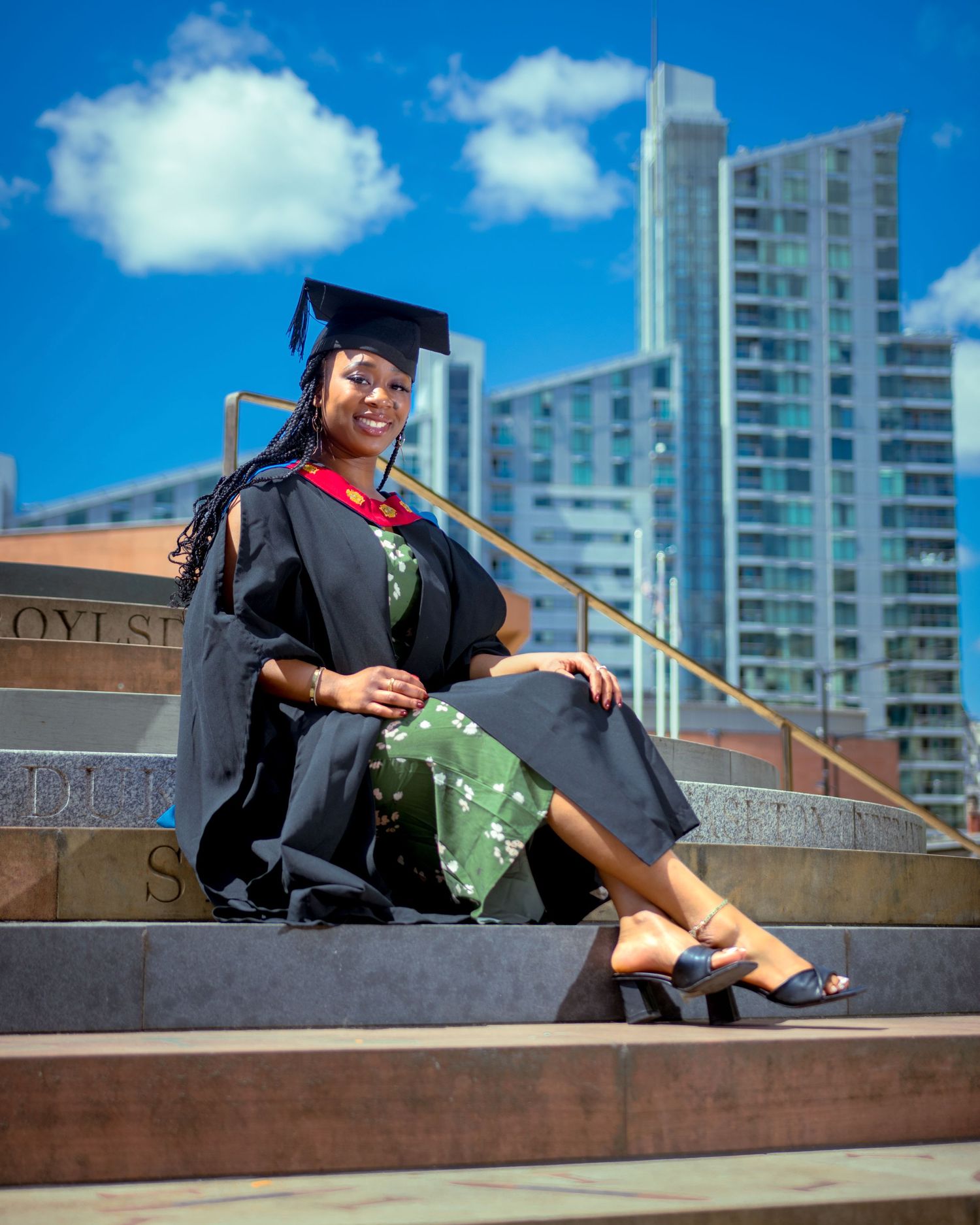 Graduate in cap and gown sitting on steps with city skyline backdrop.