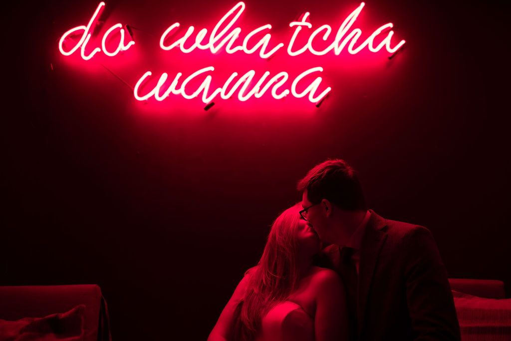 A groom kisses the bride under a neon sign that reads: Do whatcha wanna.