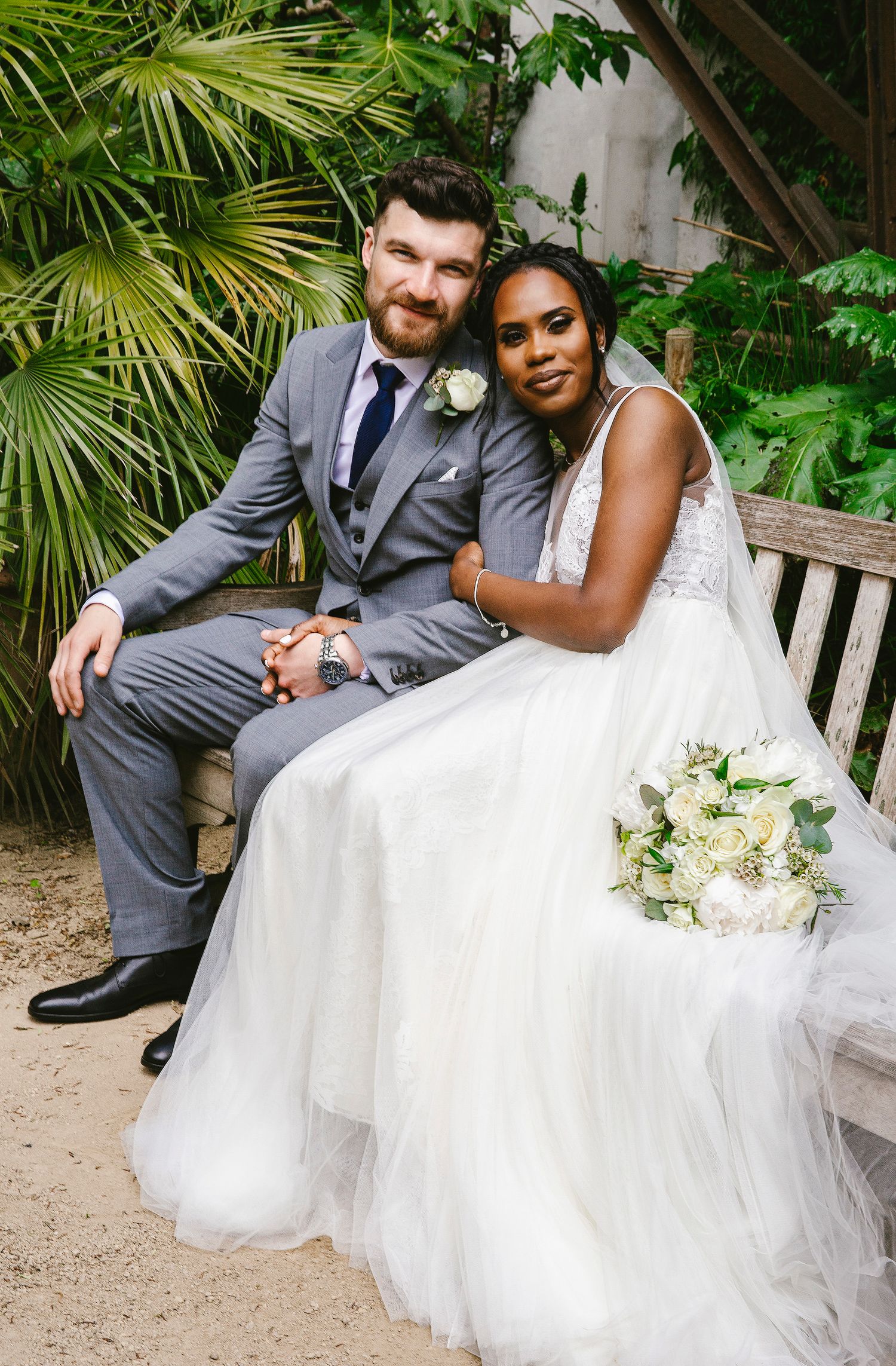 A wedding couple sits on a wooden bench surrounded by lush tropical foliage and palm leaves.
