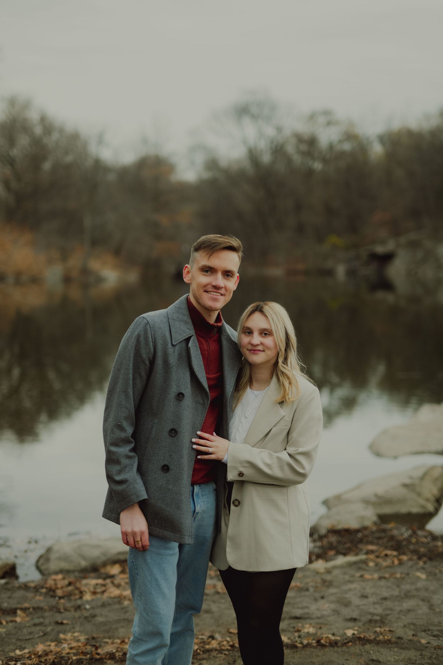 A couple shares a tender moment by a lake on an overcast autumn day with city skyline in background.