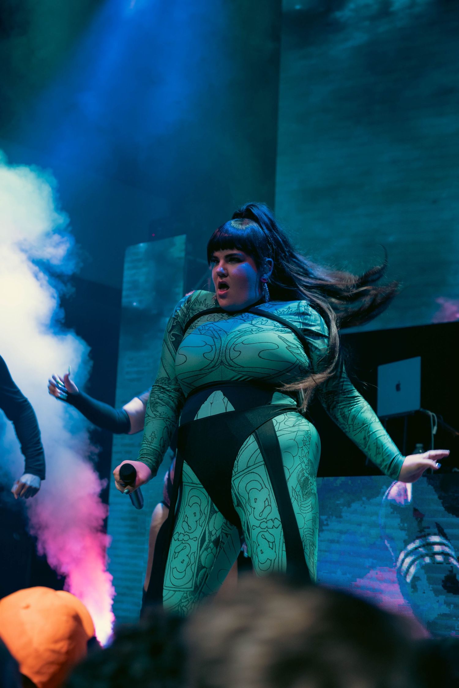 Performer in green patterned bodysuit dances dramatically on stage with colorful smoke effects in the background.