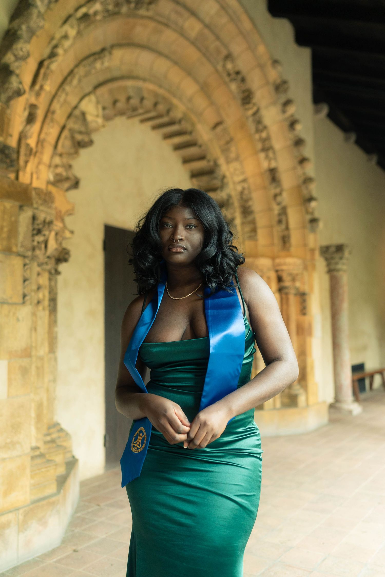 Person in green dress and blue vest poses against ornate stone archway architecture.