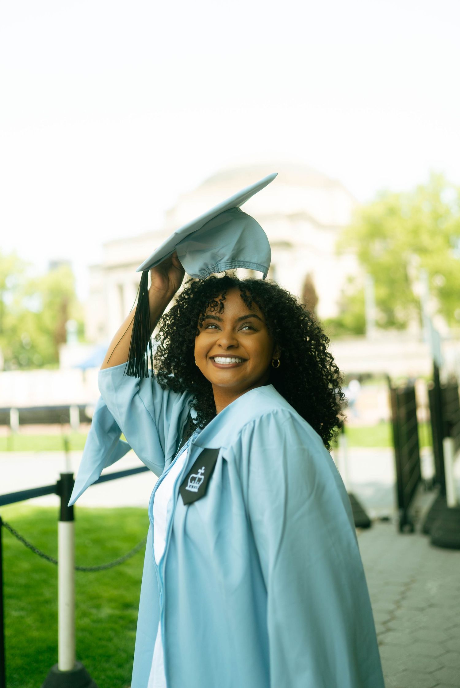 Graduate in light blue gown celebrates outdoors on a sunny day.